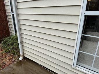 Beige siding on a building with a white window frame and a downspout. A concrete sidewalk is in the foreground.