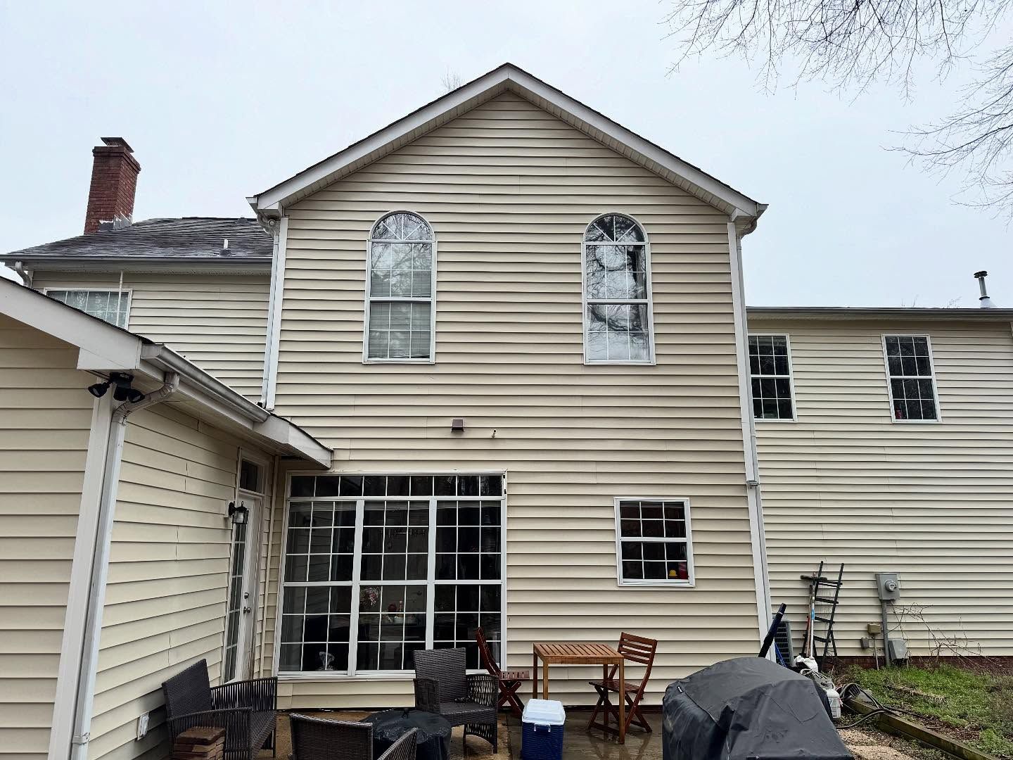 Back of a light-colored house with a large windowed sunroom, two arched windows, and a brick chimney on a cloudy day.