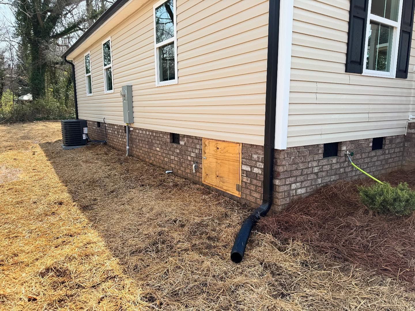 Beige house with brown brick foundation and black trim. A black downspout and vent are visible.