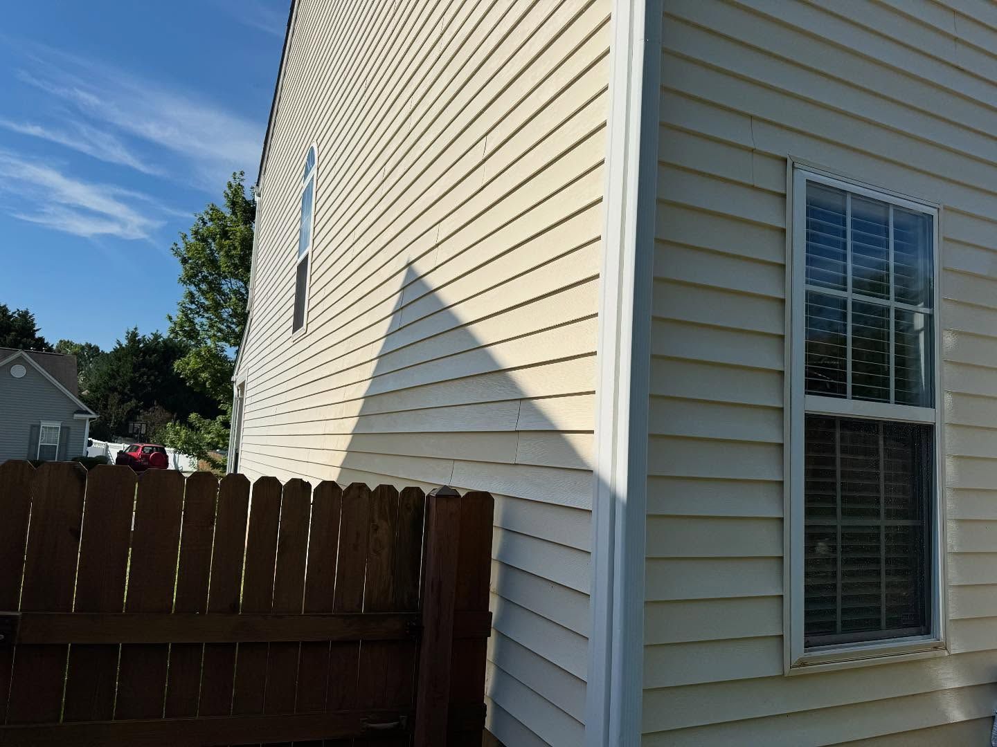 Beige siding of a house with a fence and a window on a sunny day, with a shadow cast across a portion of the wall.
