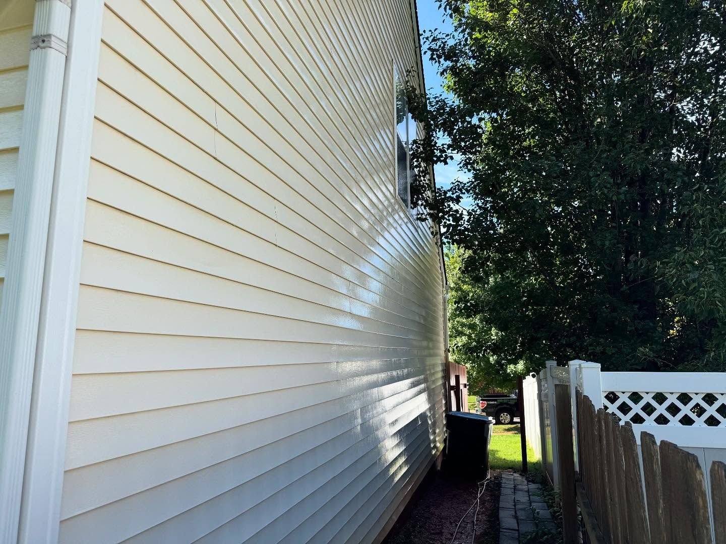 Side of a light yellow house with white trim. A narrow walkway separates the house from a wooden fence and trees.