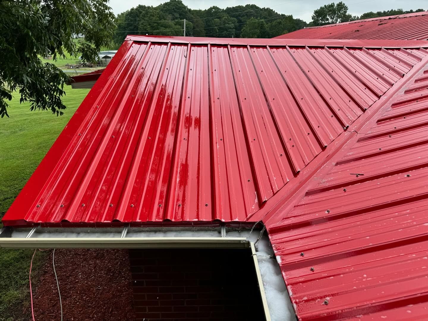 Red metal roof on a building, viewed from the side, with a white gutter. Green grass and trees in the background.