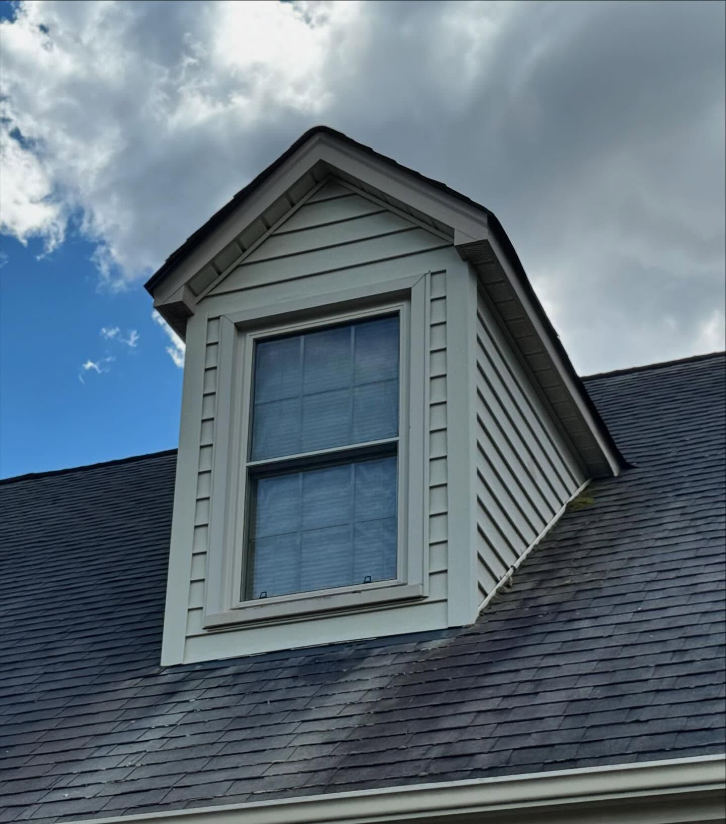 Dormer window with light-colored siding and a dark shingled roof against a cloudy sky.