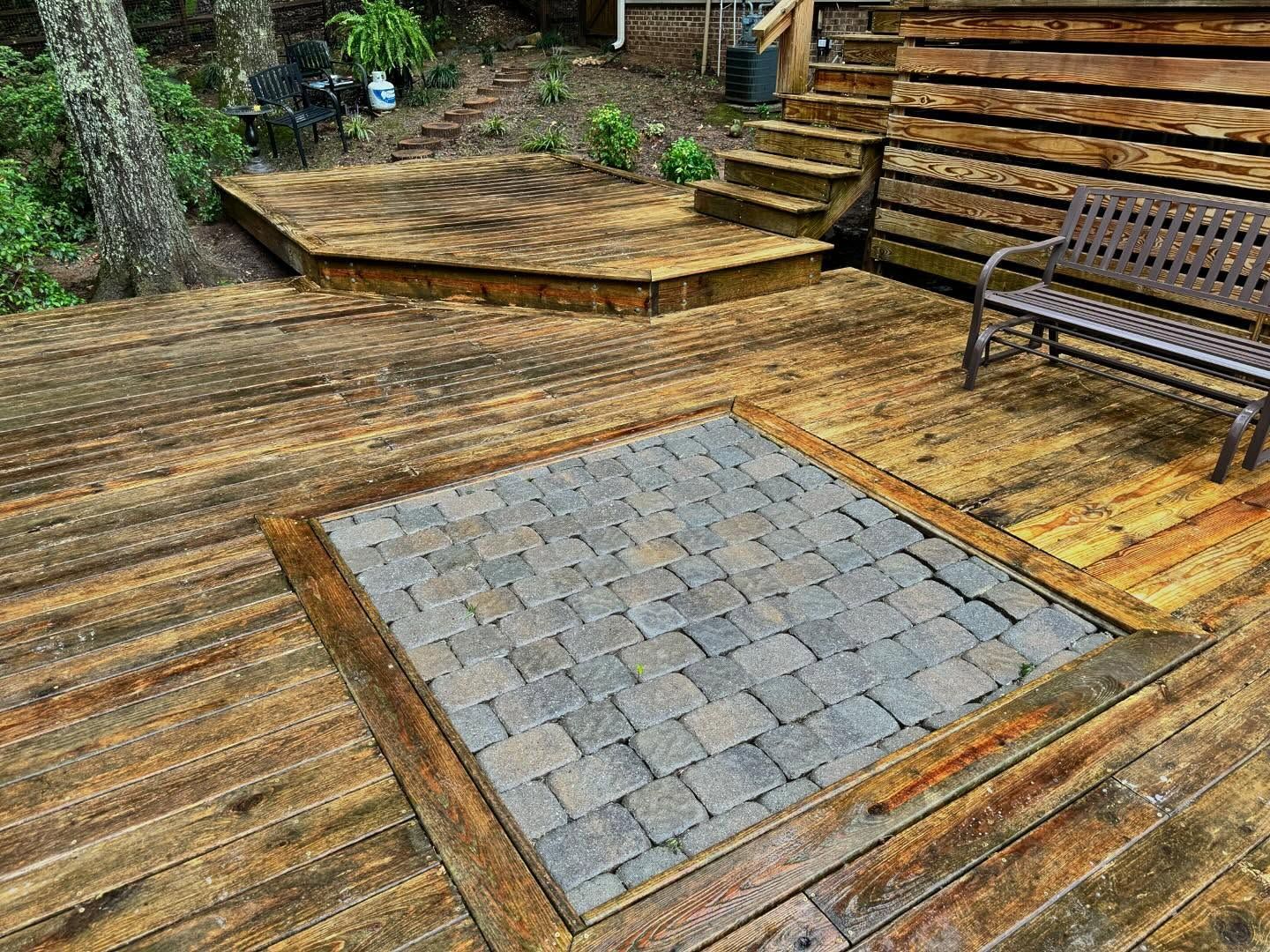 Wooden deck with a square stone patio inset, steps, and a bench. The deck is surrounded by foliage.