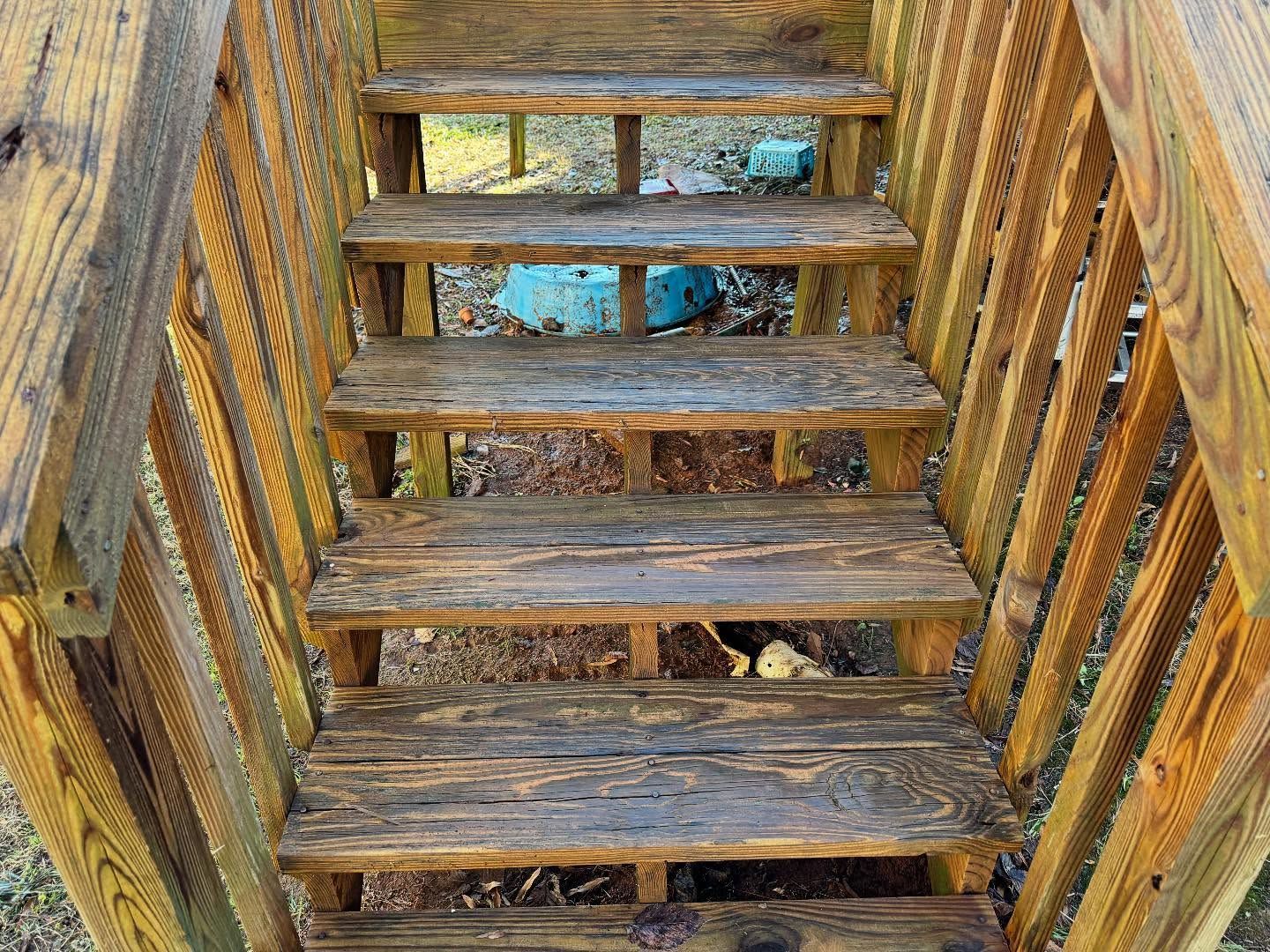 Wooden outdoor staircase, weathered and worn, leading upwards. Rails and steps are brown.
