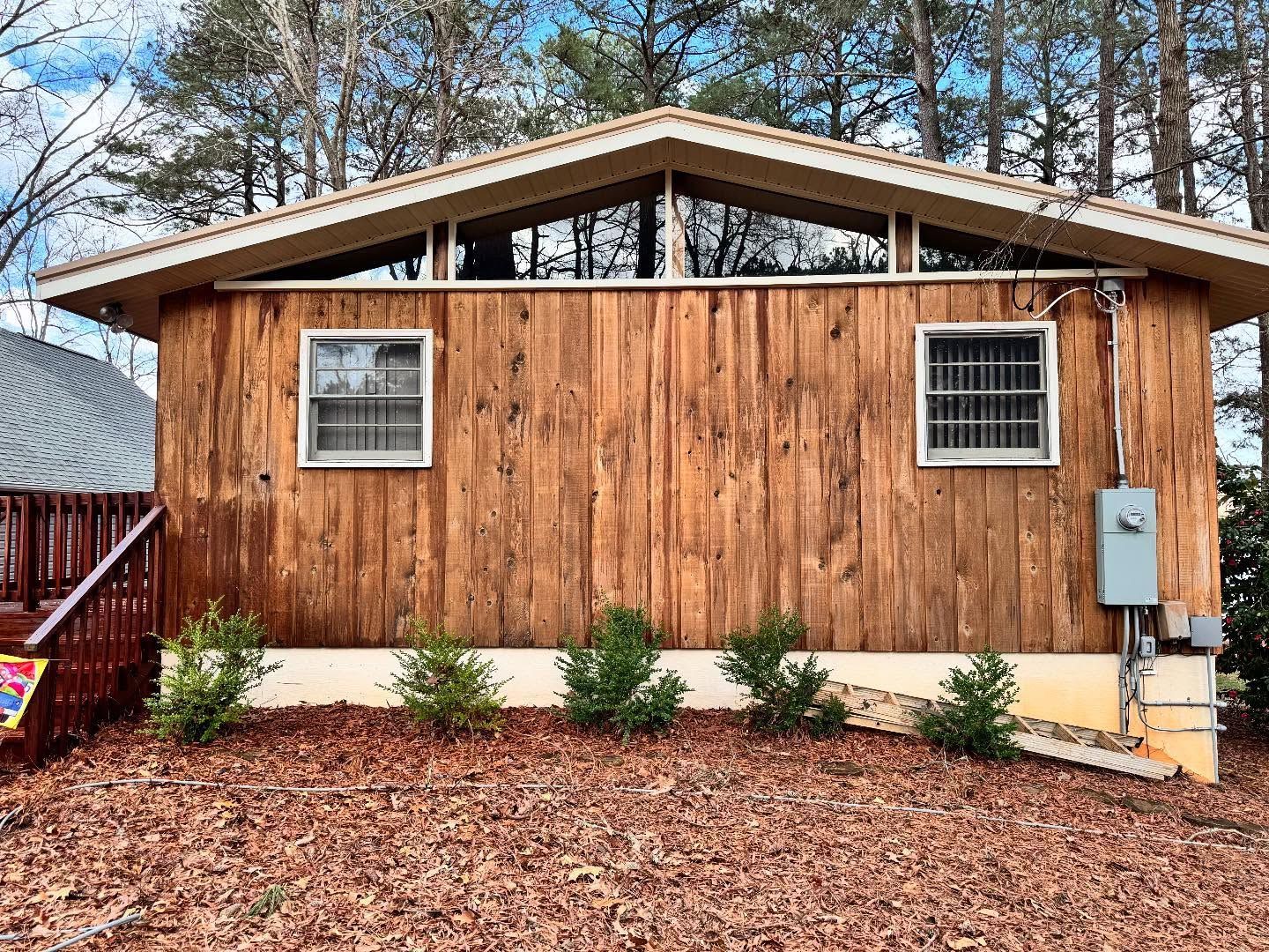 Wooden cabin with two windows and angled upper windows, in a wooded setting. Green shrubbery lines the base.