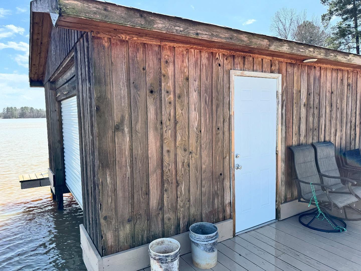 Wooden boathouse on the water with white door and closed garage door. Two chairs and buckets sit on the wooden deck.