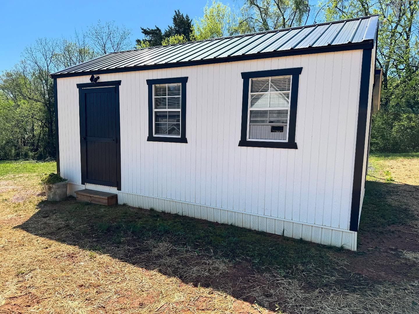 White shed with black trim, a black door, two windows, and a metal roof, set in a grassy area with trees in the background.