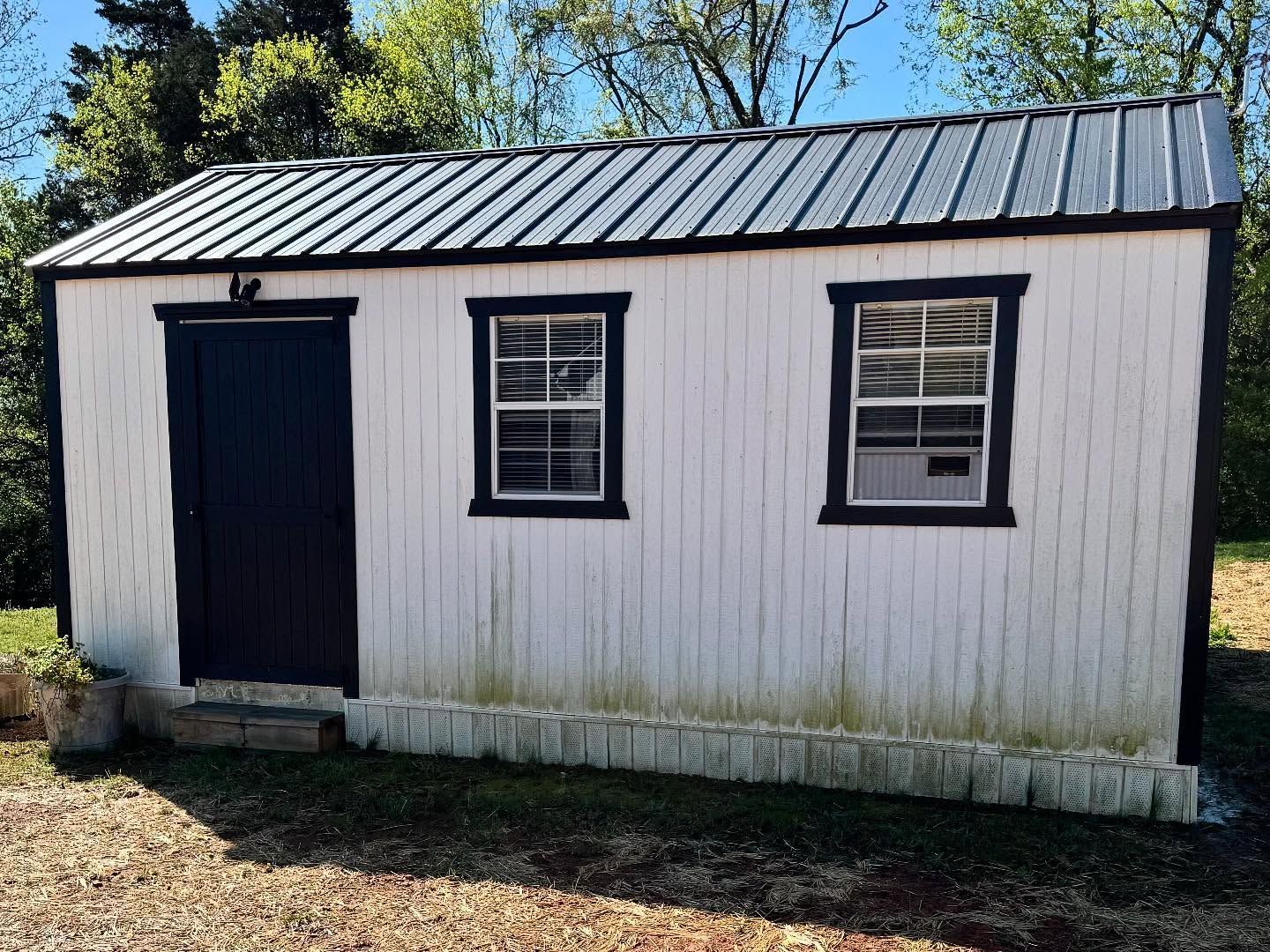 A white shed with a black door, window frames, and roof trim. The shed has a metal roof and two windows.
