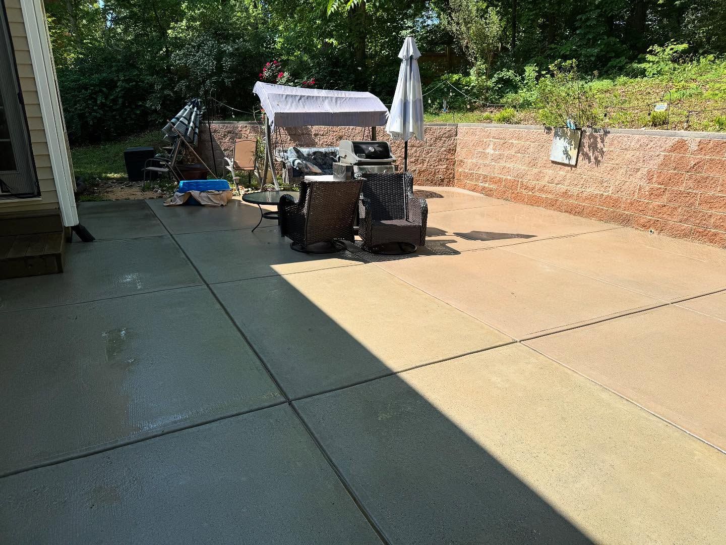 A sunny patio with a table, chairs, and an umbrella. A raised brick wall is on the right, and trees are in the background.