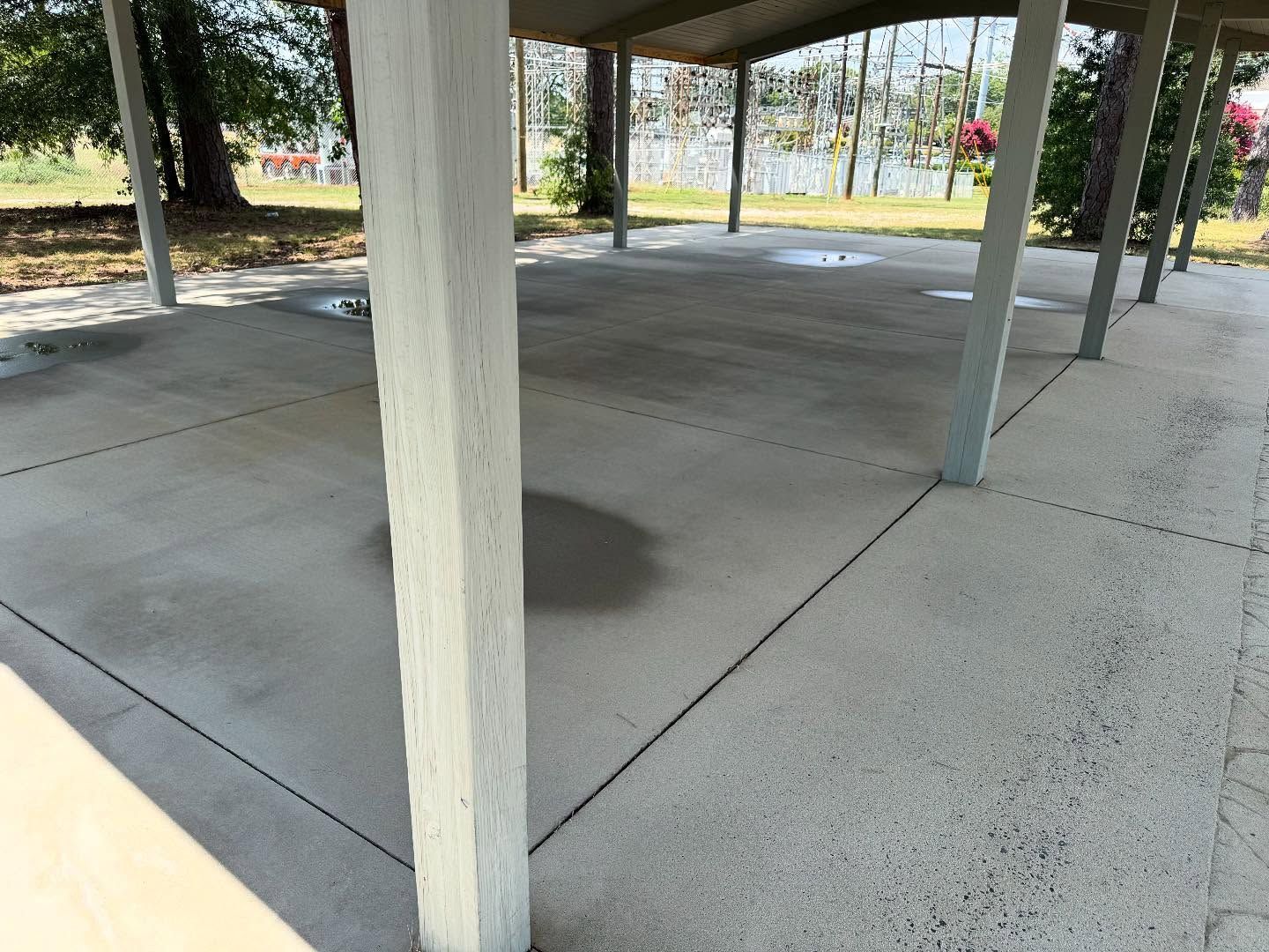 Concrete pavilion with gray pillars, dappled sunlight, and a view of trees and water in the distance.