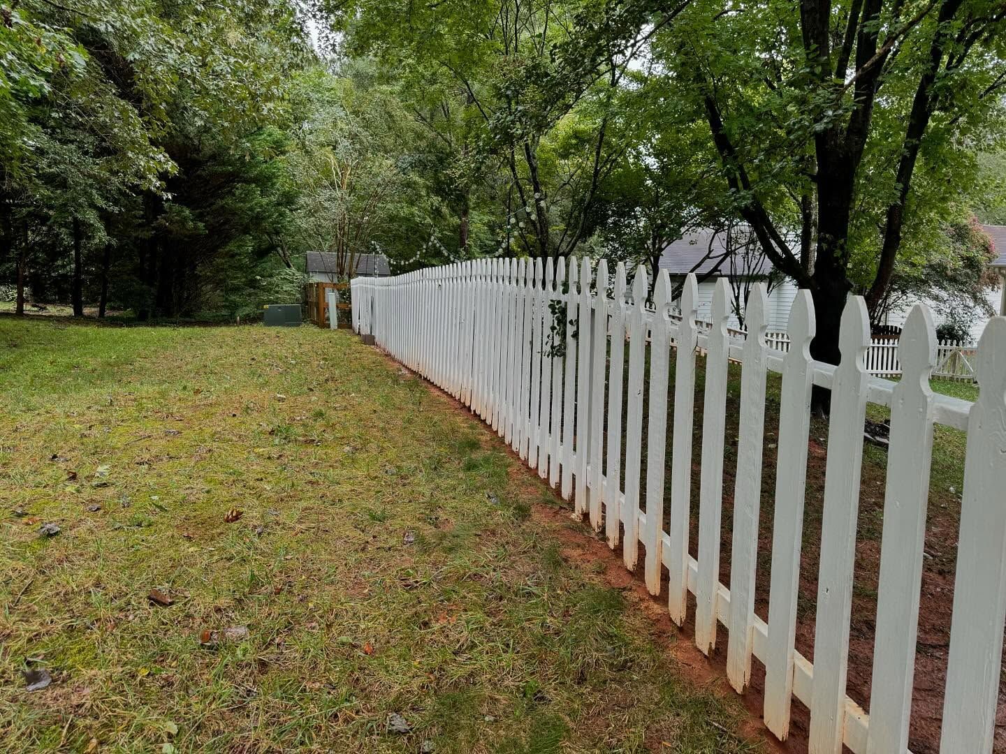 White picket fence curving across a grassy yard, with trees in the background on a cloudy day.
