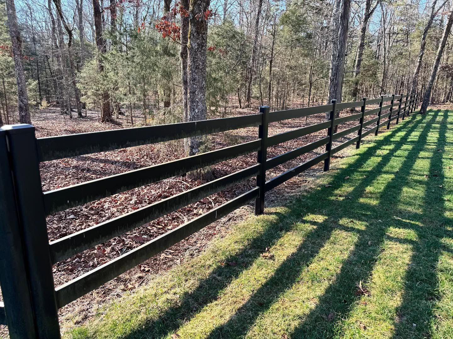 Black wooden fence in a grassy area with long shadows cast by the sun. Trees in the background.