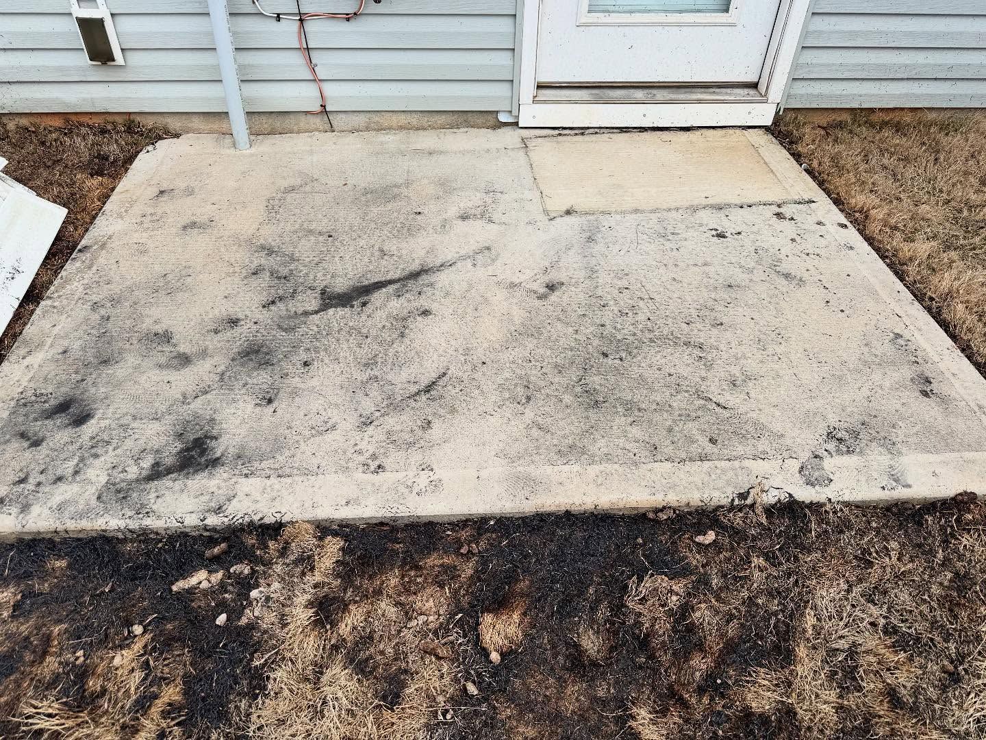 Concrete patio with black marks, next to a door with a small welcome mat. Brown grass surrounds it.