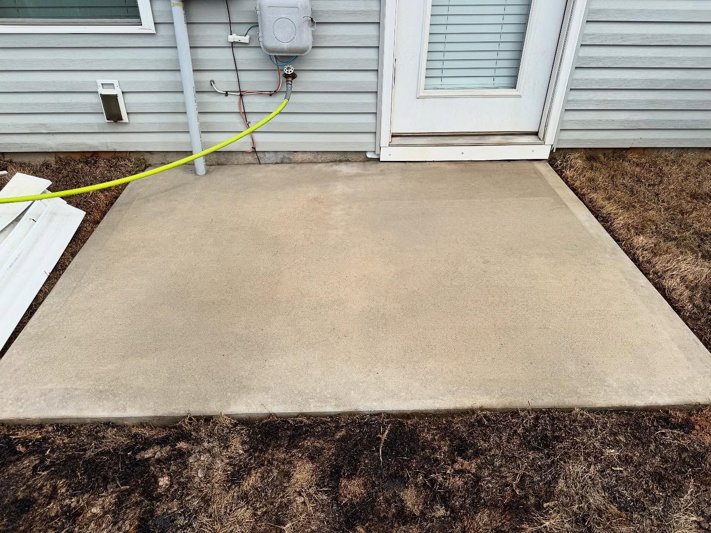 A concrete patio outside a house with a door. A gas meter and green hose are mounted on the siding above the patio.