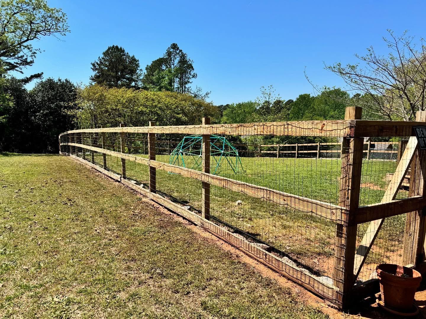A wooden fence with a wire mesh encloses a grassy area under a bright blue sky. A wooden gate is on the right side.