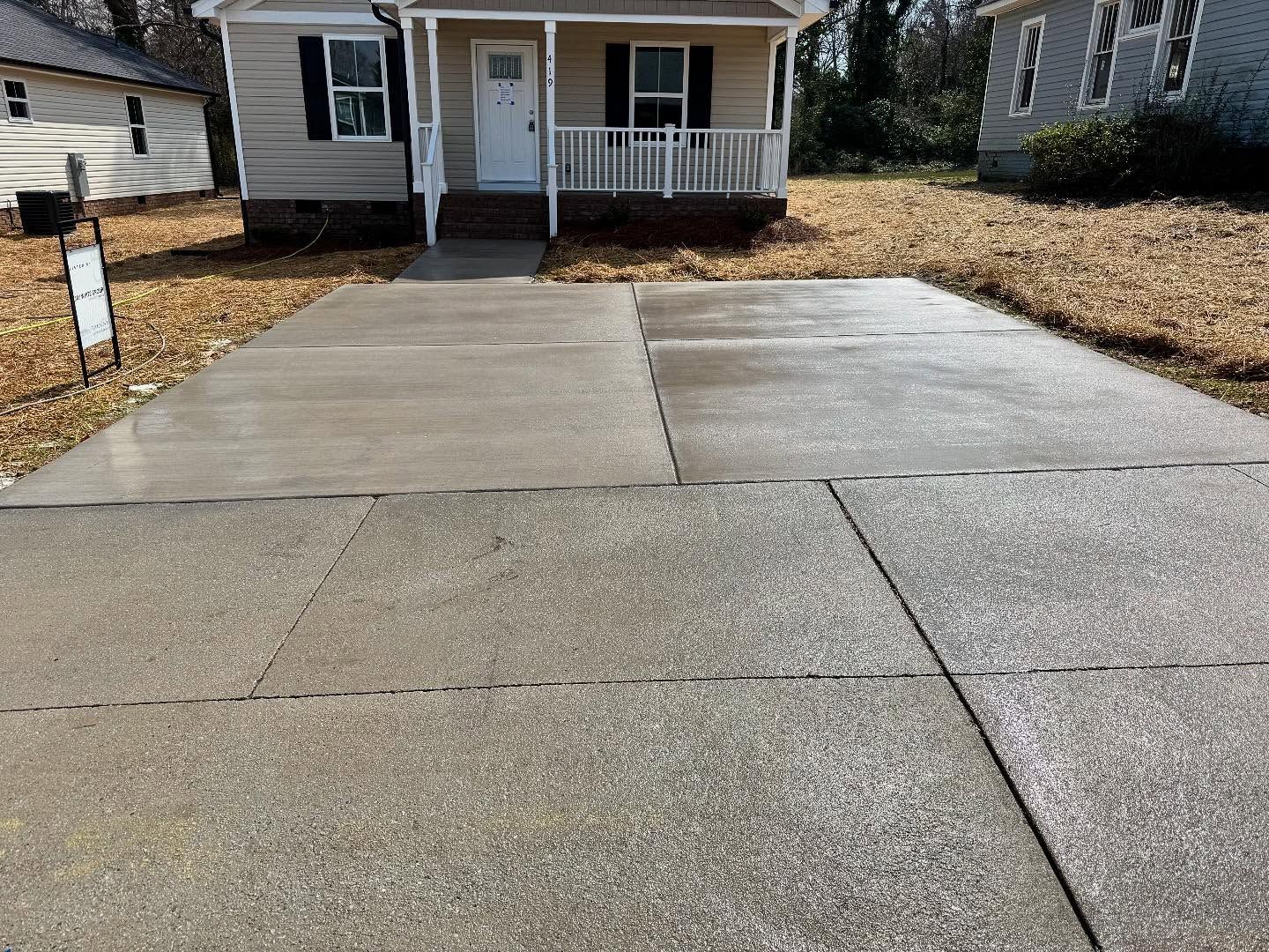 Driveway leading to a small house with a porch. The driveway is made of concrete and is slightly wet.