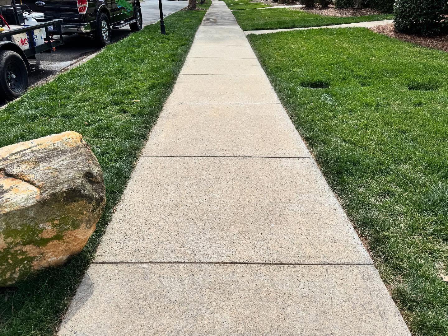 Sidewalk bordered by green grass, with a large rock in the foreground, and a truck visible in the background.