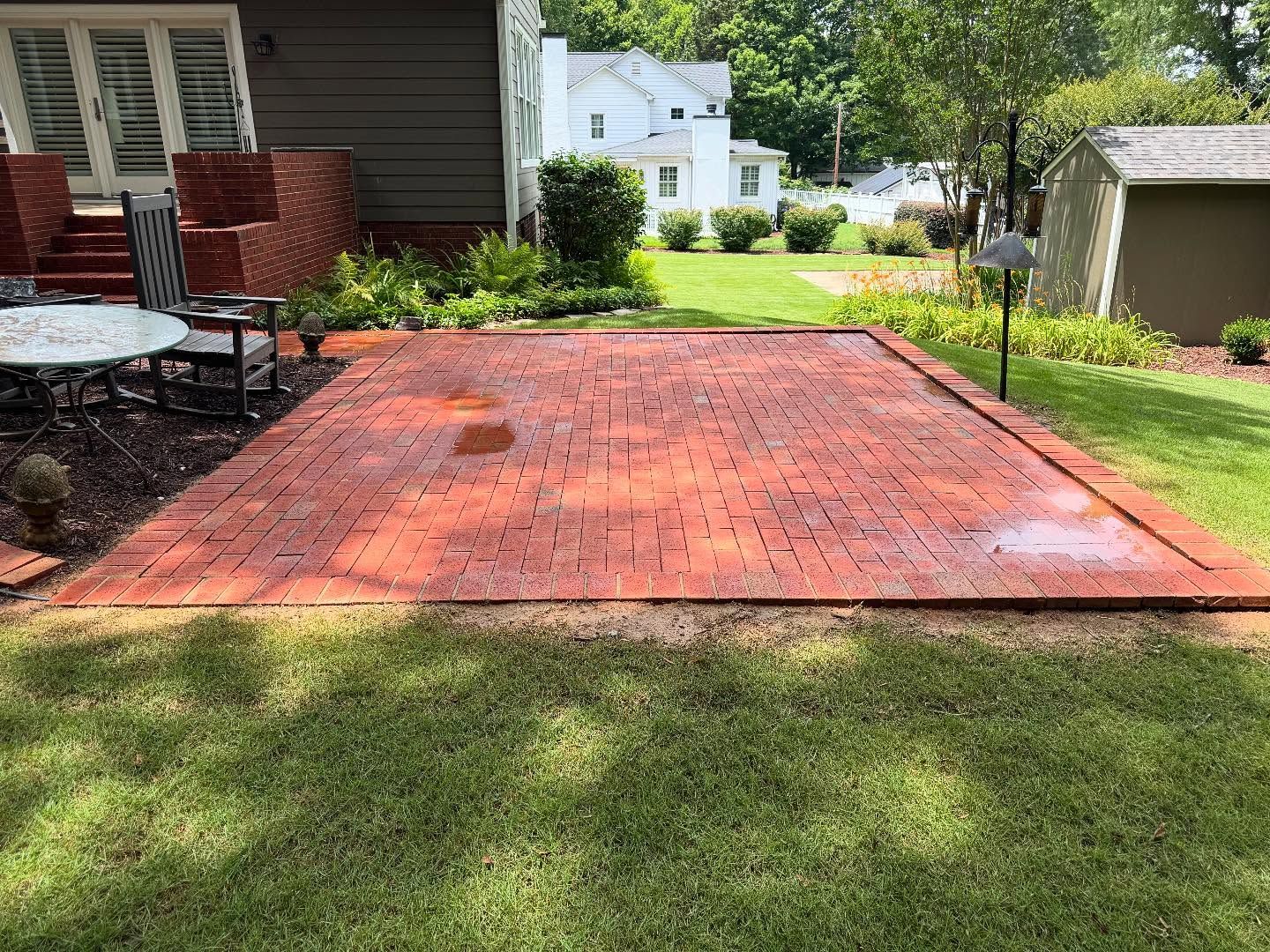 Brick patio in a grassy yard, bordered by a brick edge. The patio is wet, with a house and other buildings in the background.