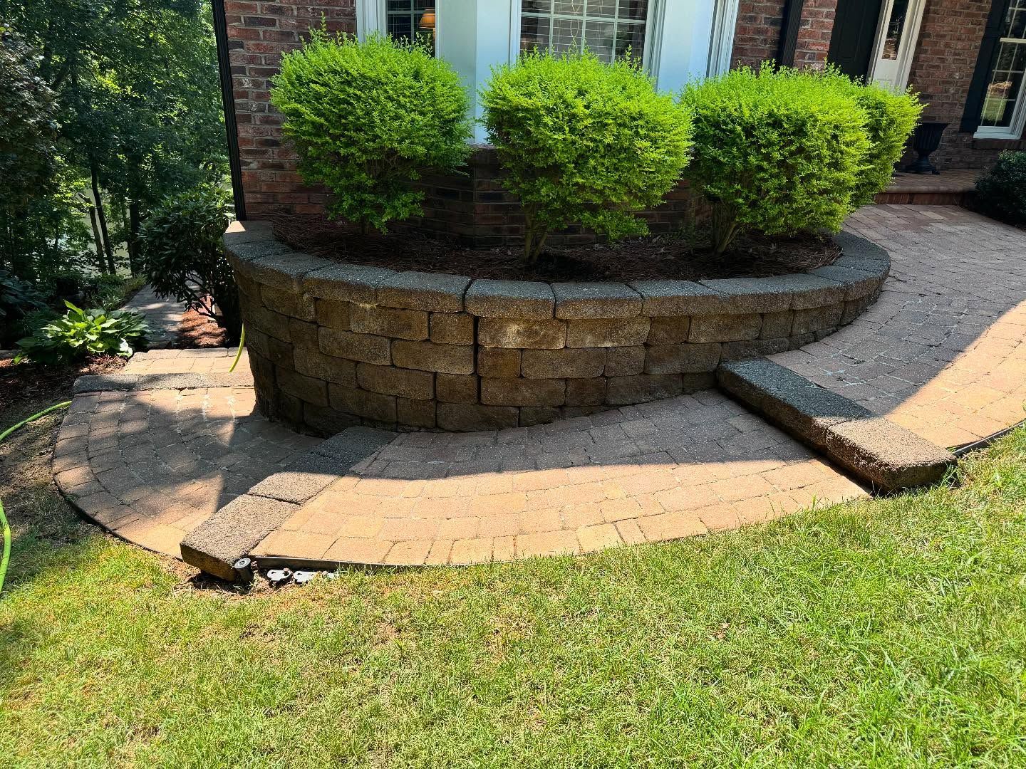 Circular brick retaining wall holding green bushes, with a stone path leading up to it, in front of a brick house.