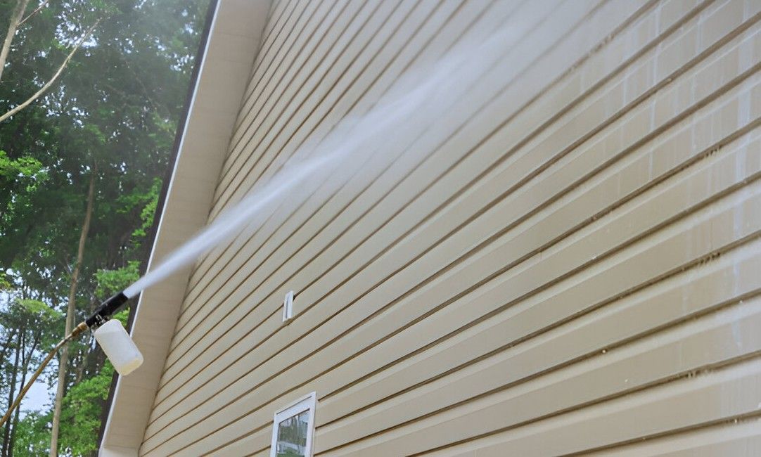 A person power washing tan siding of a house near a window with white trim. Water sprays from the nozzle.