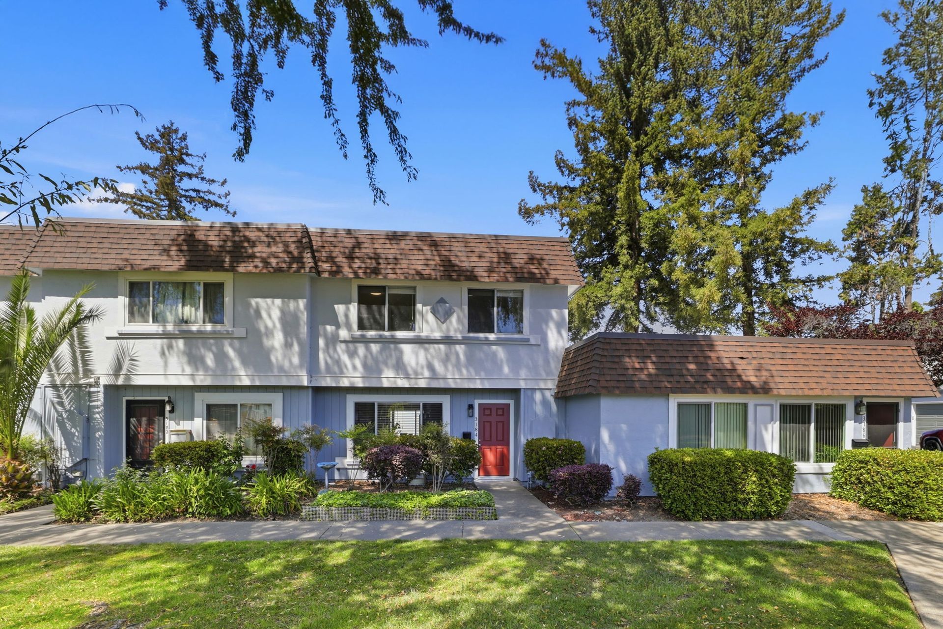 Two-story white townhouse with brown roof, red door, and manicured front lawn under a clear blue sky