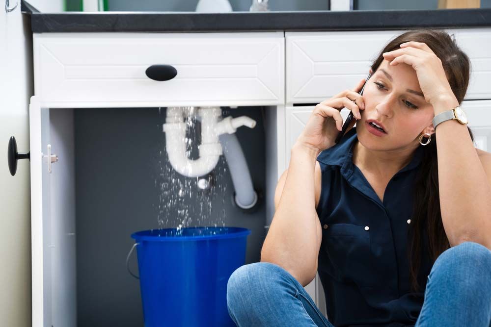 A Woman Is Sitting On The Floor Under A Sink Talking On A Cell Phone — Tweed Heads Leak Detection in Casuarina, NSW