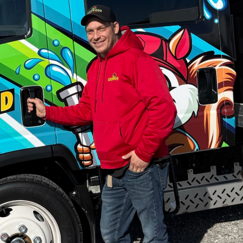 A man wearing a red shirt and a black hat is standing in front of a pine tree.