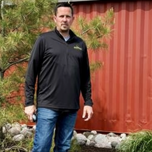 A man wearing a redwood top is smiling in front of a red wall.