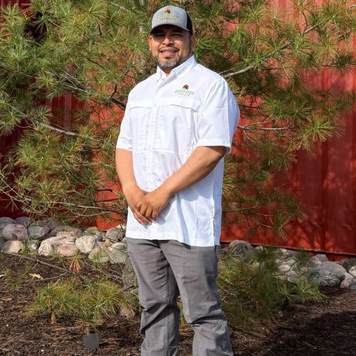 Tish A woman in a white polo shirt is standing in front of a pine tree.