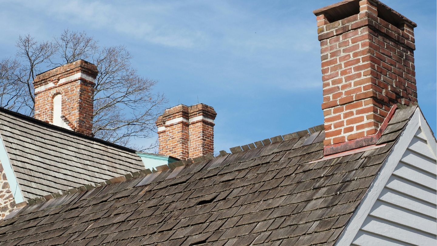 Three brick chimneys on a weathered wooden shingle roof against a blue sky.