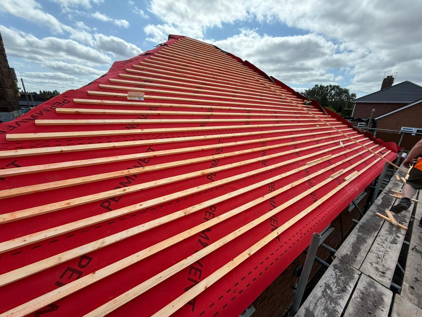 Roof construction in progress; red underlayment with parallel wooden battens, blue sky.