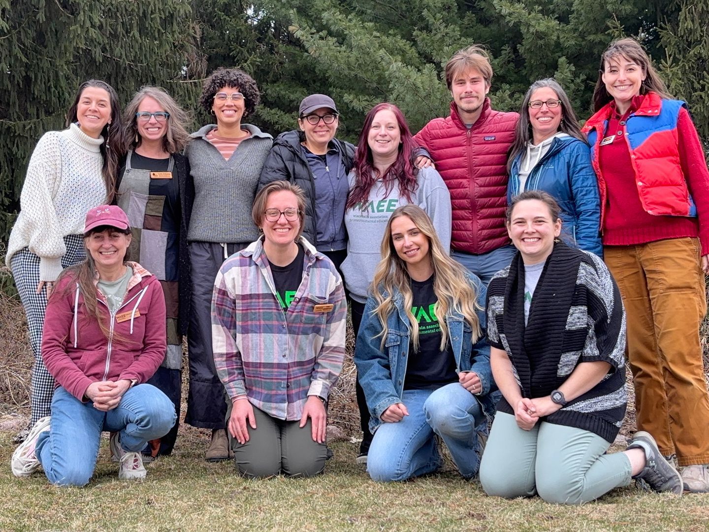 A group of 14 people smiling and posing for a photo outdoors in front of a tree line on a sunny day.