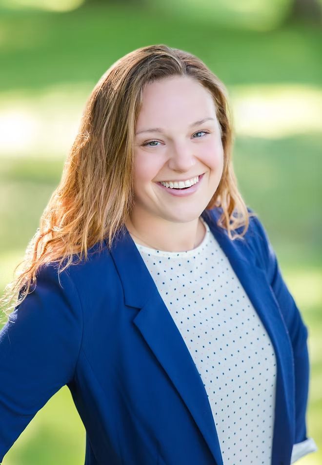 Woman smiling outdoors, wearing a blue blazer over a white polka dot blouse.