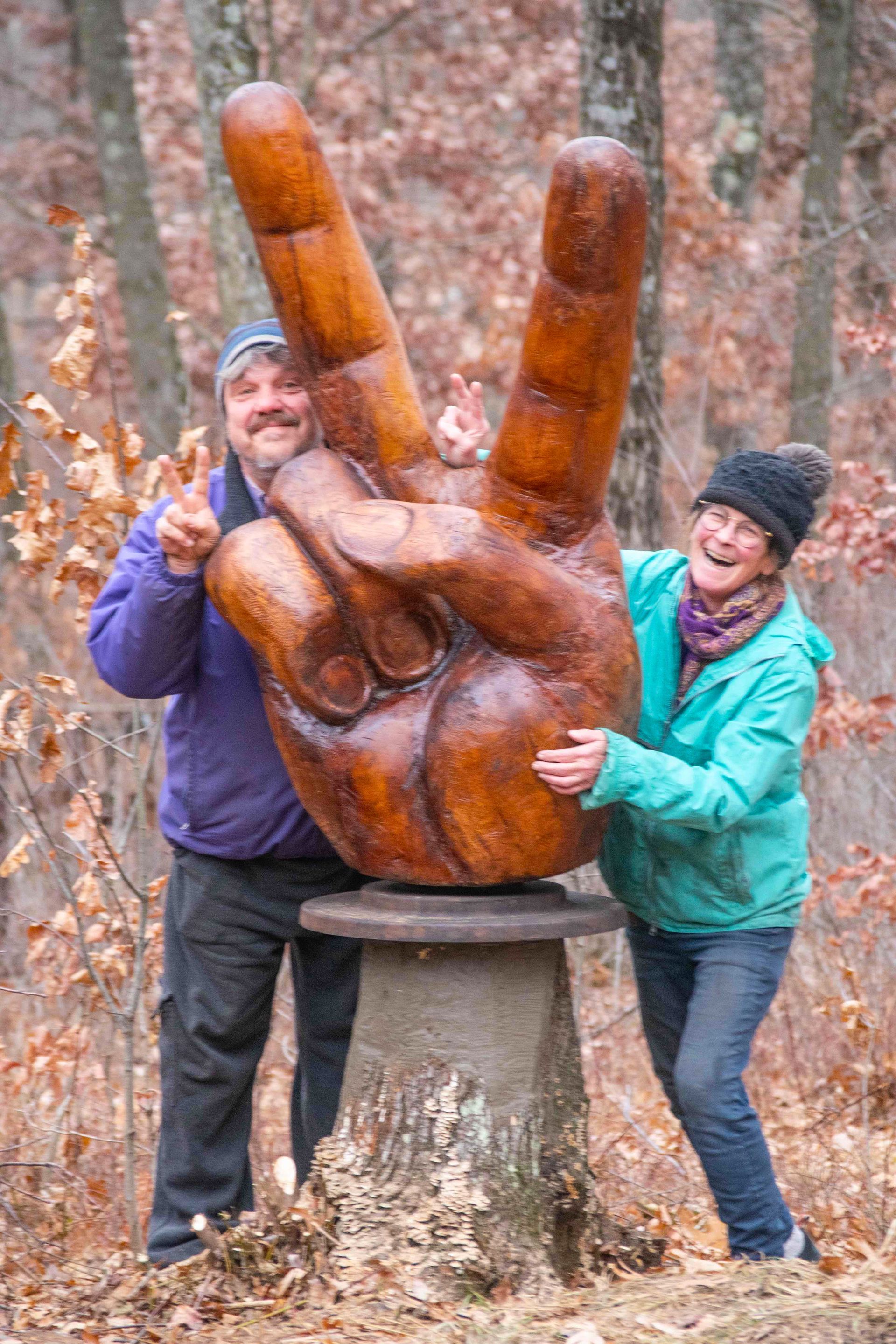 A man and a woman standing behind a large wooden carved hand showing the Peace Sign. 