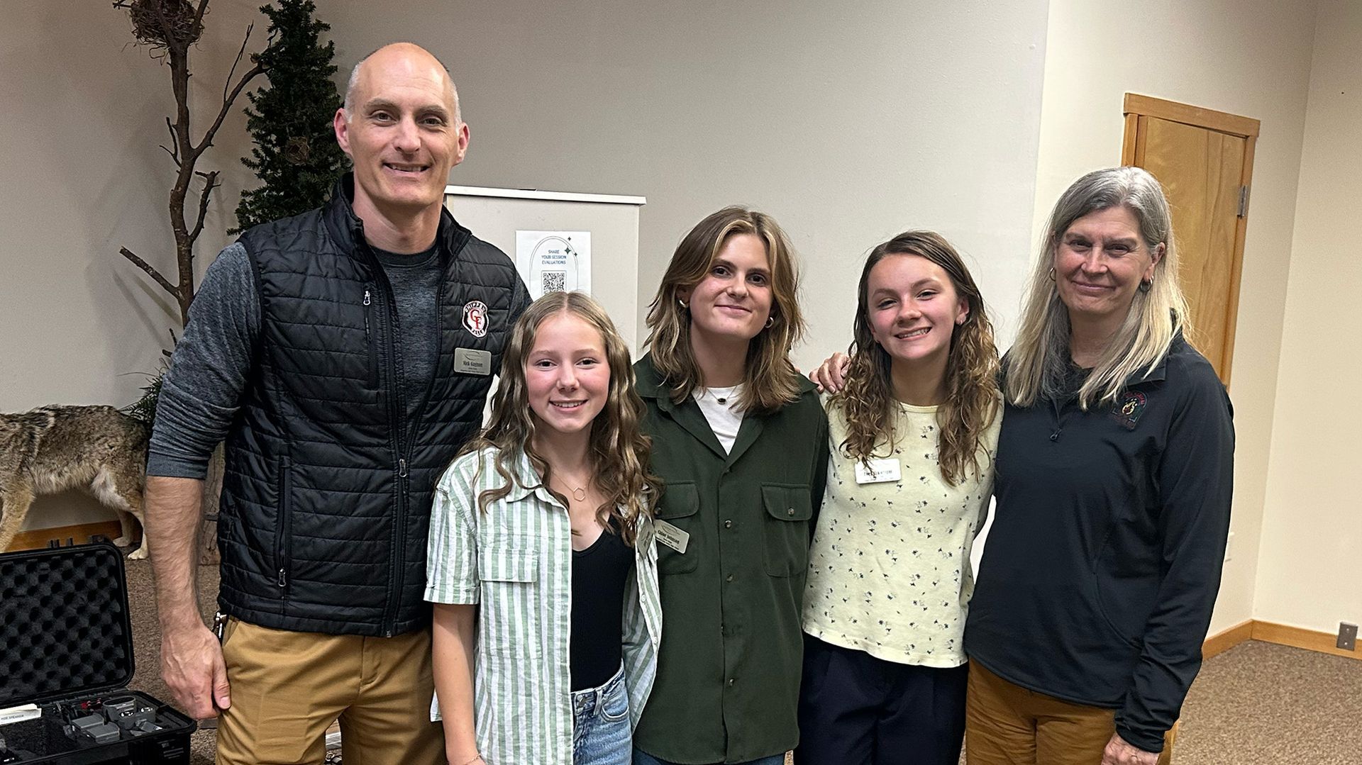Group of five people posing indoors: a man in a vest, three young people, and a woman.