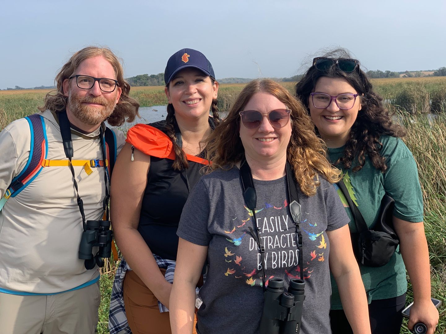 Four smiling hikers posing together in a sunny open field, with backpacks and outdoor gear.