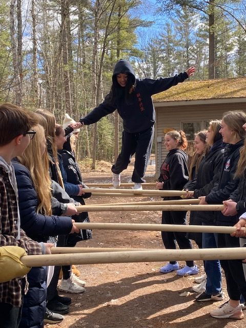 People balancing a person on poles. Outdoors. Building in background. Sunny.