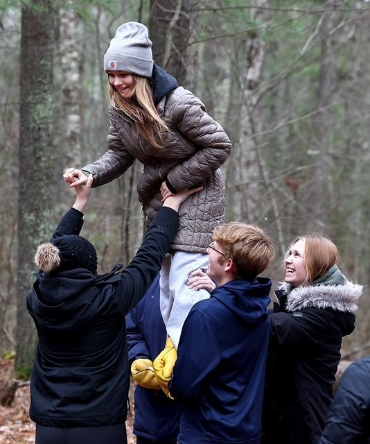 People lifting a person up in a forest; the person on top smiles, hands raised, others support her.