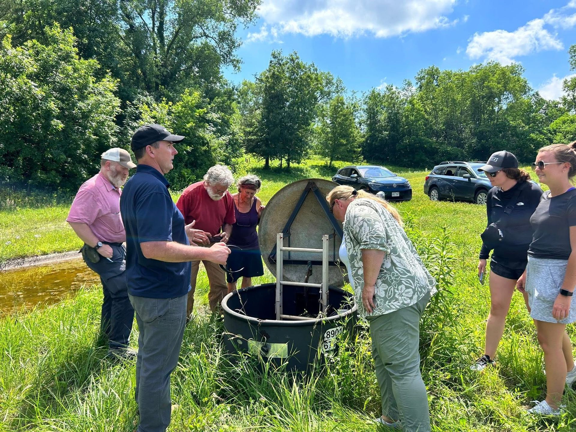 A group of people gather around an open well in a grassy field.