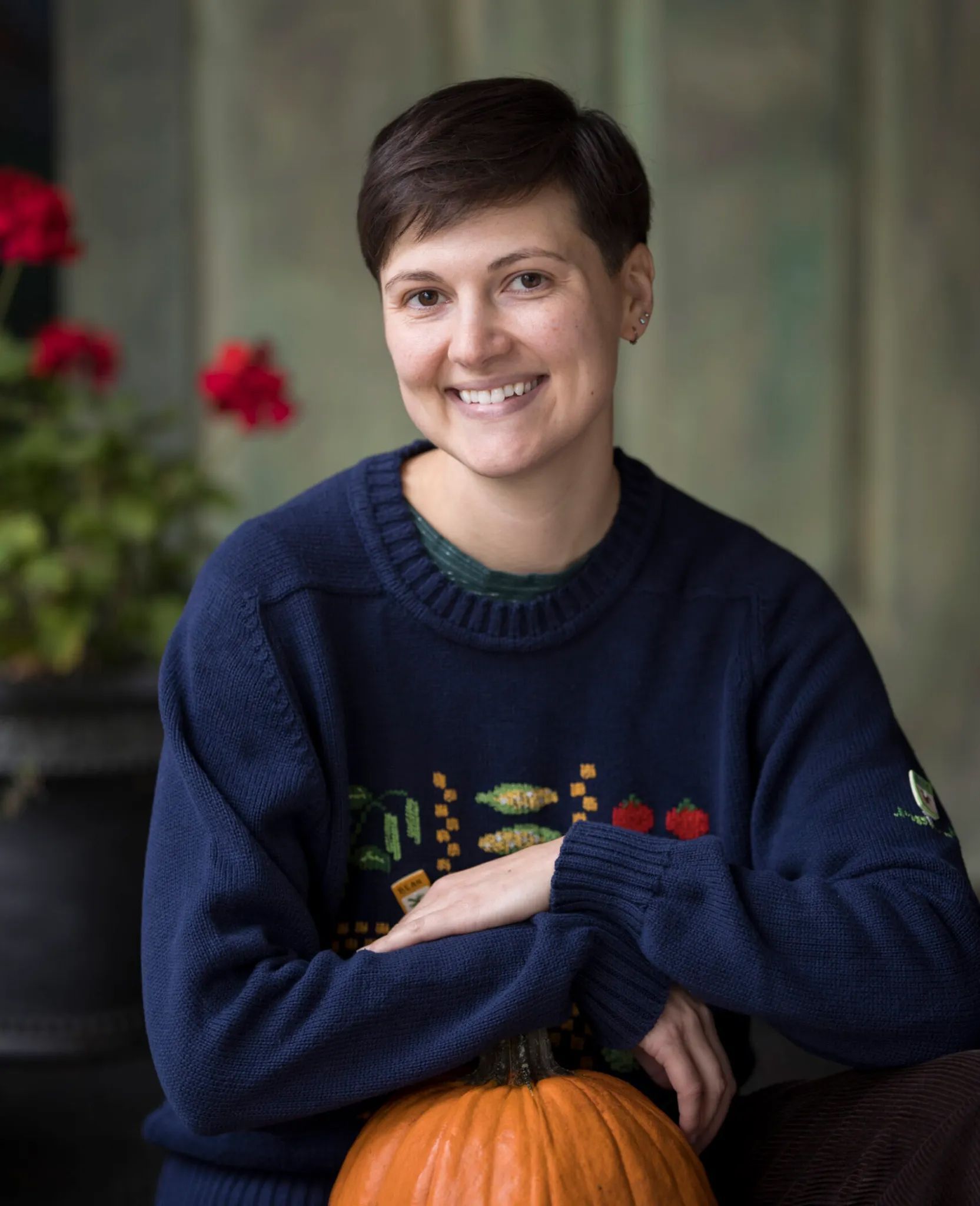 Woman in blue sweater smiles, resting arm on pumpkin, red flowers in background.