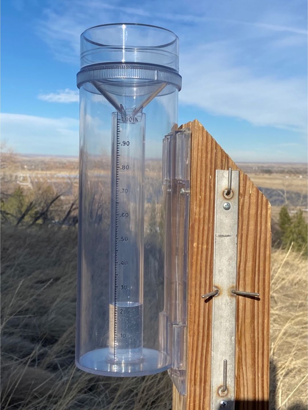 Clear rain gauge mounted on a wooden post outdoors against a blue sky and field.