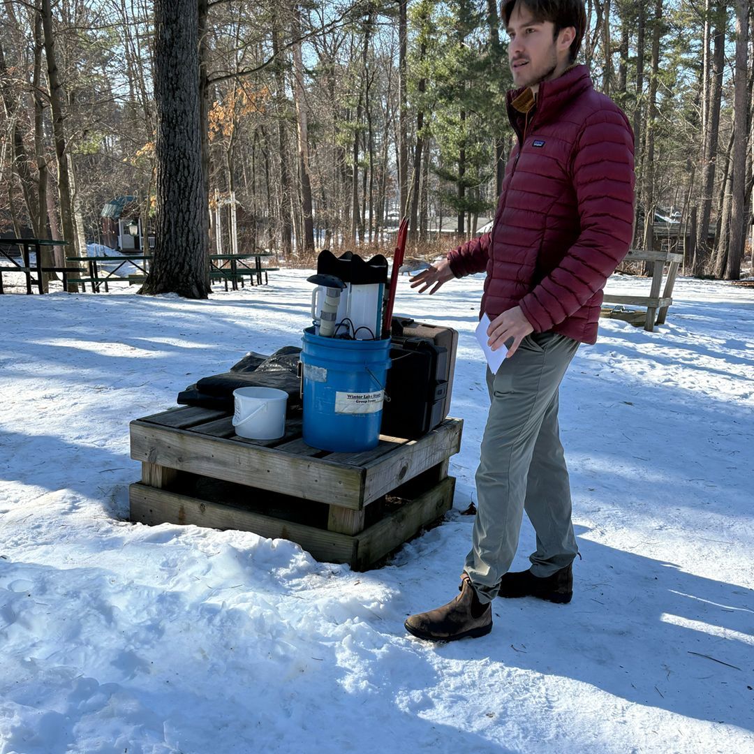 A person in a maroon puffer jacket stands by a wooden pallet topped with a blue bucket in a snowy, wooded park setting.