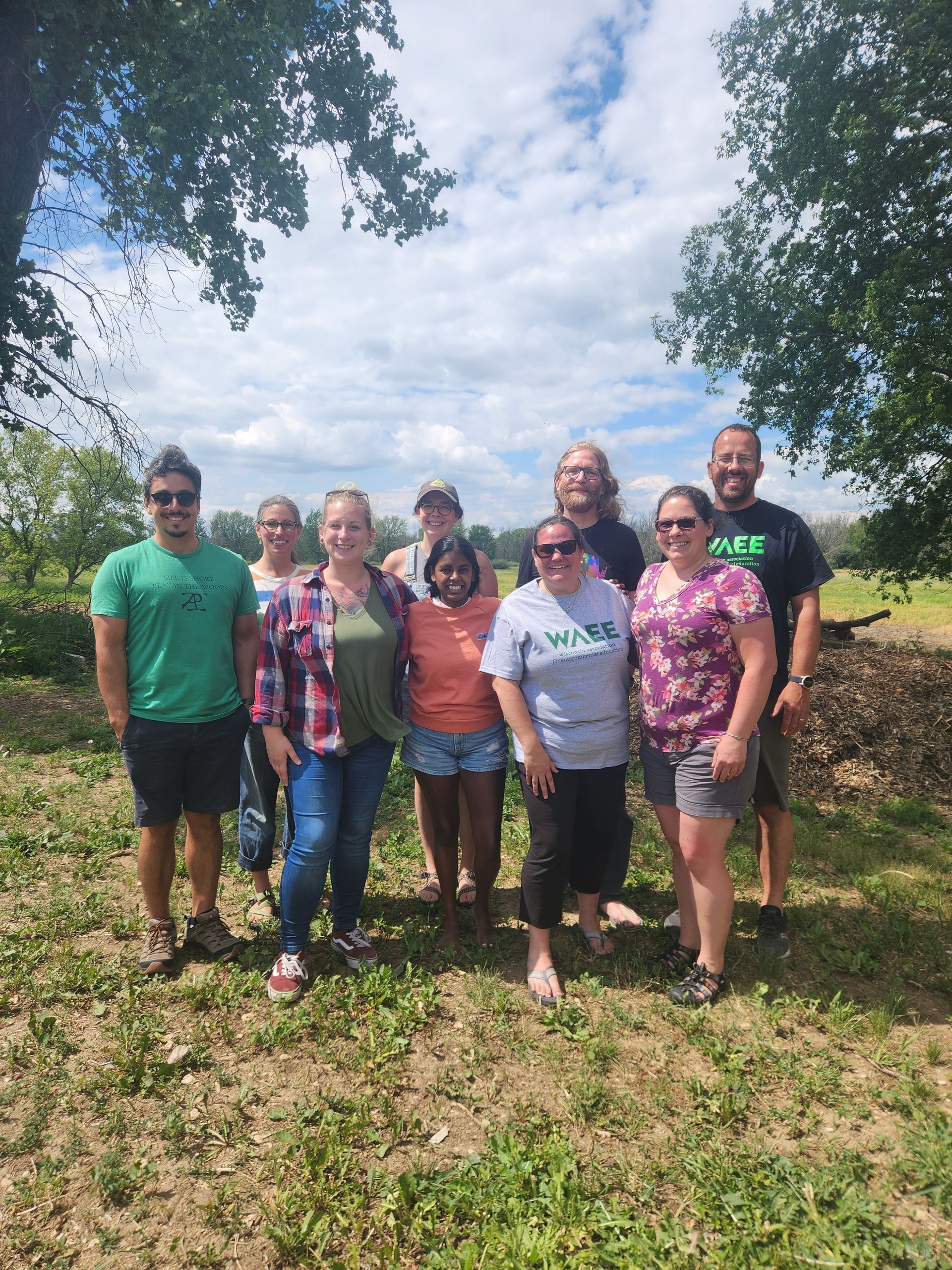 Group of people standing outdoors, smiling. Sunny day, green grass, trees.