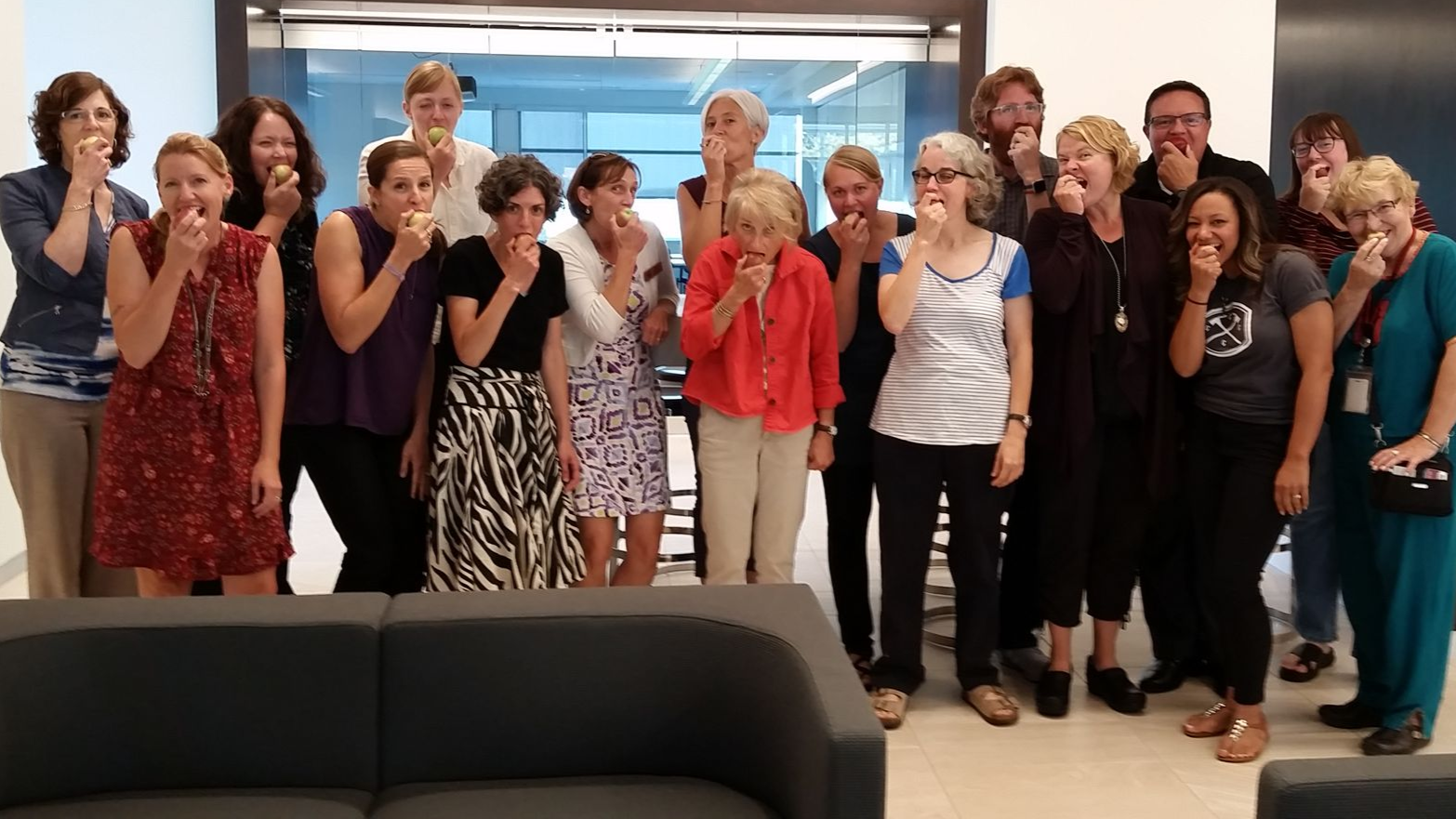 Group of people smiling with one hand holding an apple in front of their mouths, standing in front of windows and a gray couch.