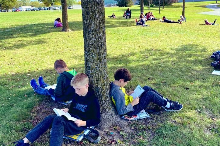 Elementary students sitting outside with backs against a tree reading.