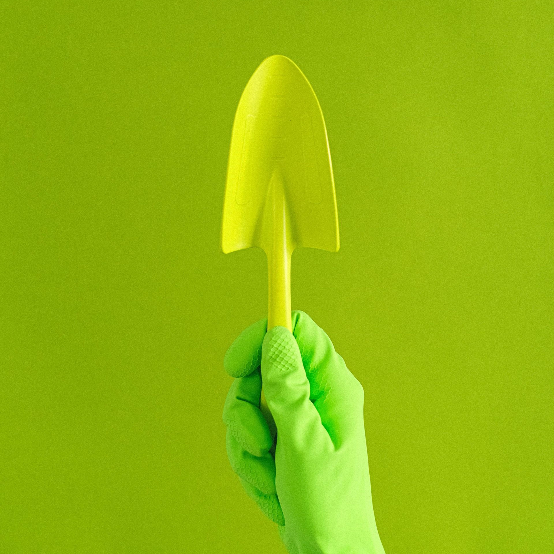Green-gloved hand holding a yellow gardening trowel against a green backdrop.