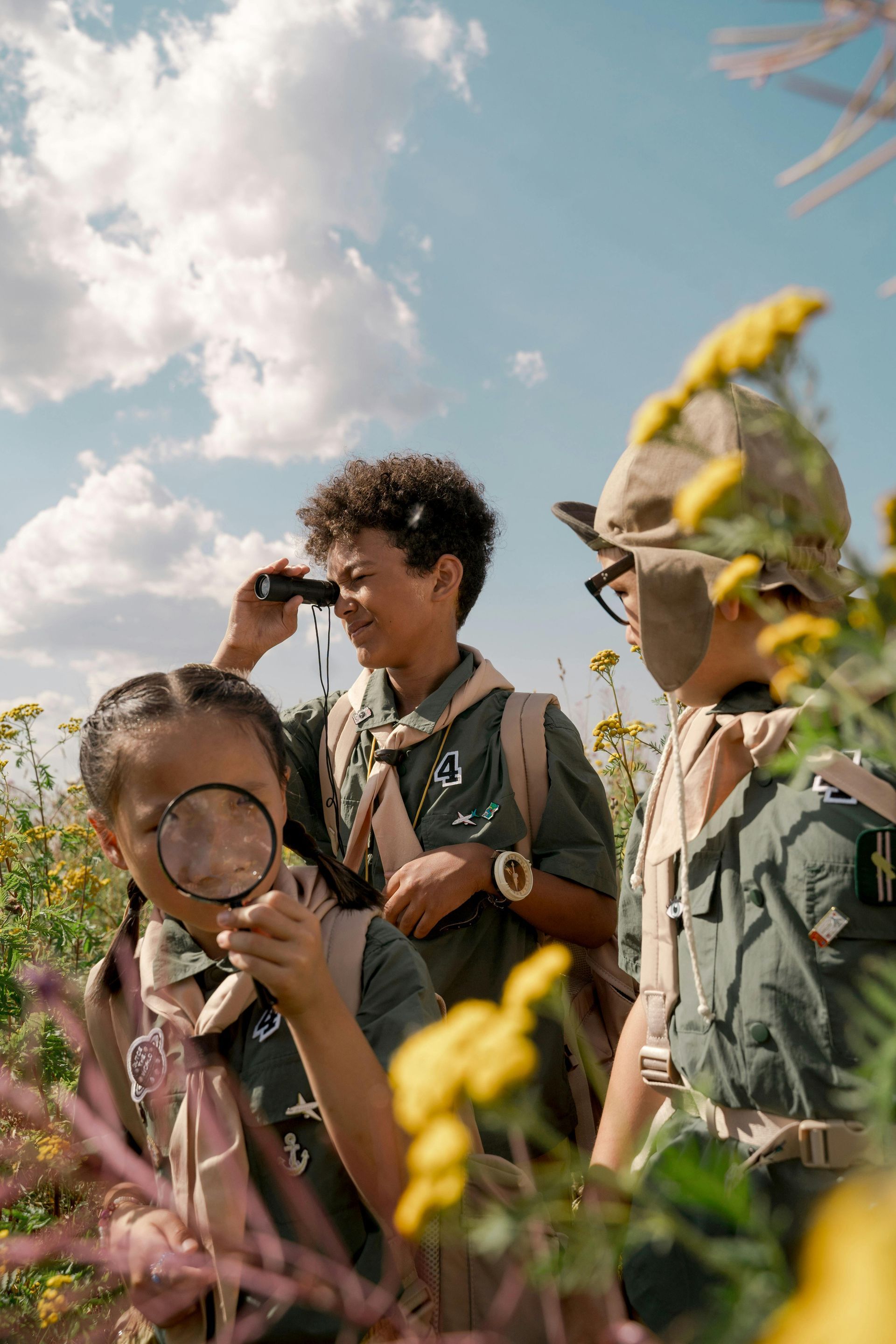 three kids outdoors in a prairie using binoculars and magnifying glasses.