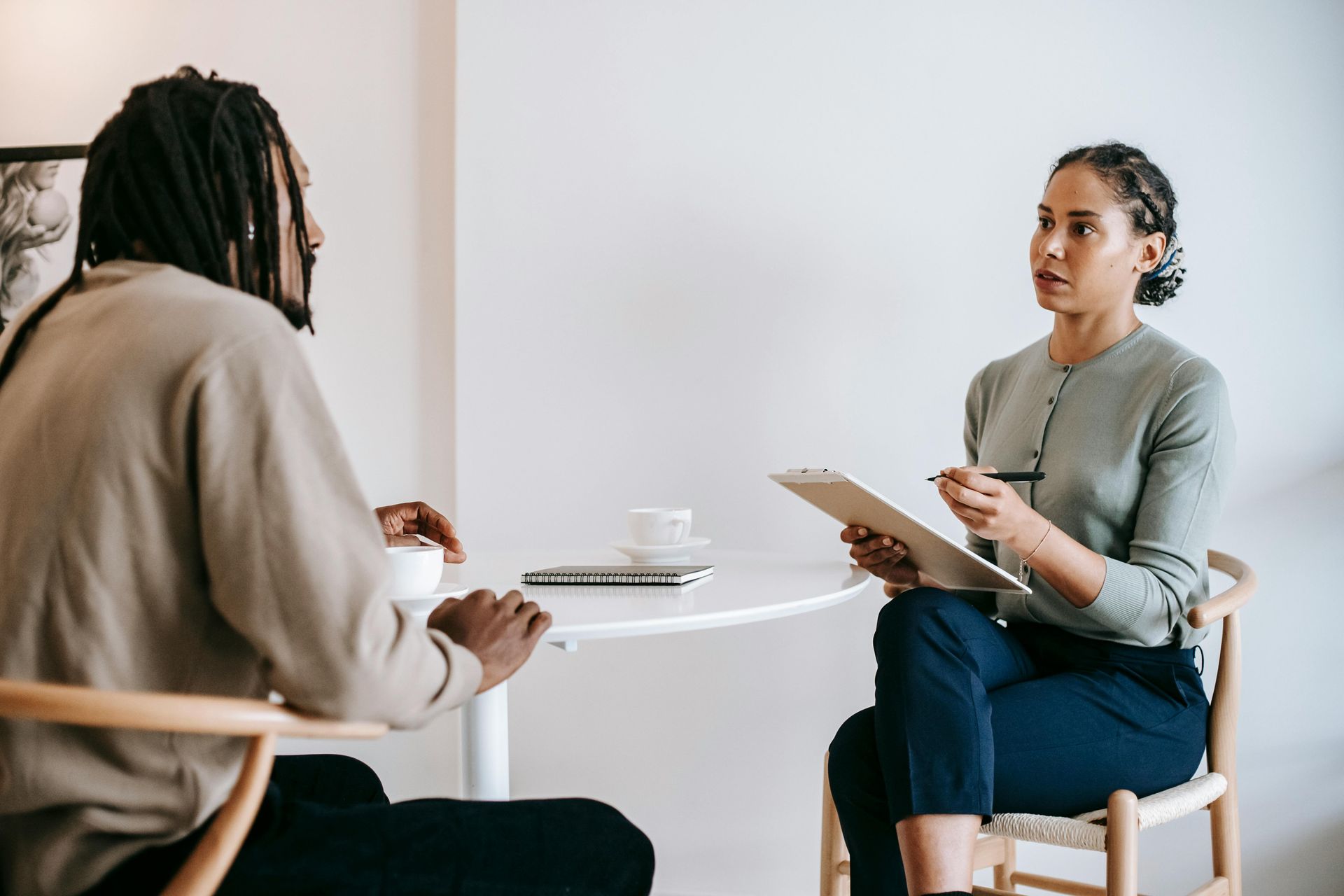 Person being interviewed by another person, both seated at a table. Woman holds clipboard and pen.