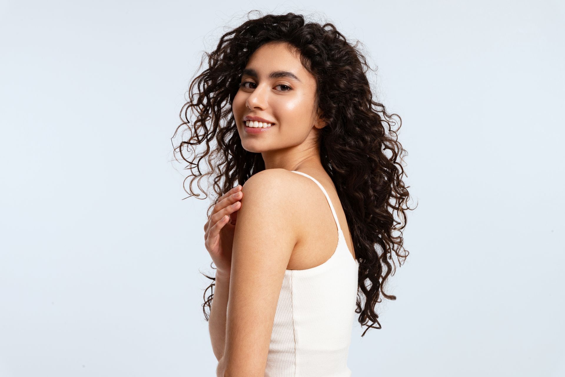 Woman with curly hair smiles, turns towards the camera, and touches her shoulder; light blue background.